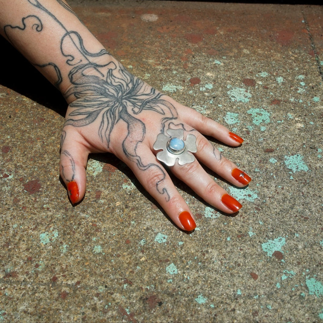 Hand with tattoos and red nail polish on a textured surface with sterling silver flower ring