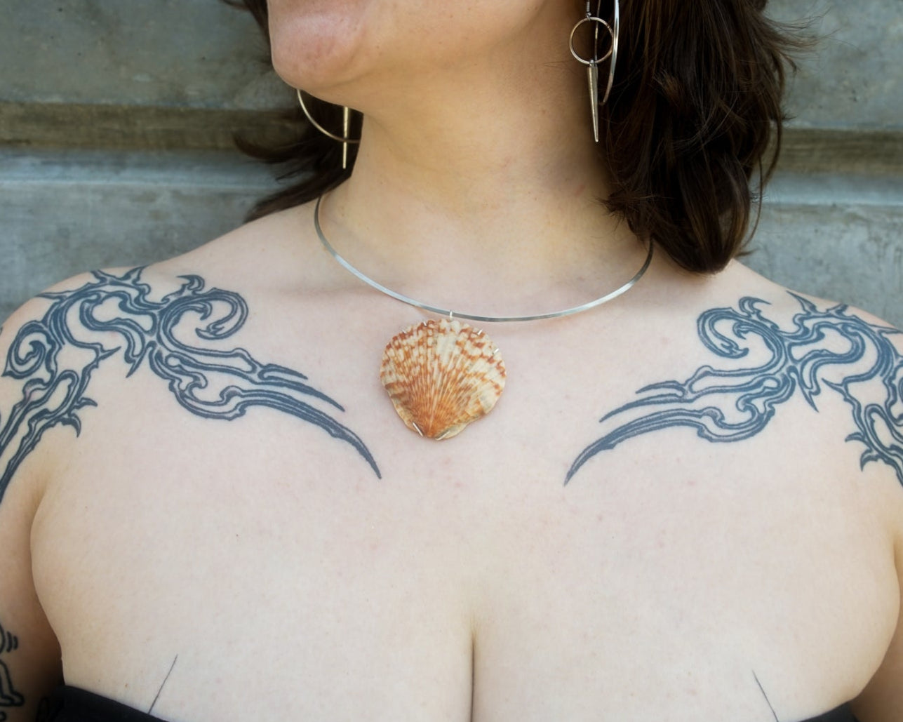 Person with tattoos on arms and chest wearing a shell necklace against a concrete wall.